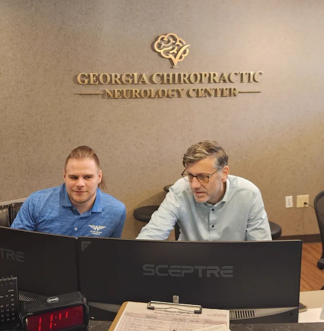 Two men work at dual Sceptre monitors at a clinic front desk. A wall sign reads “Georgia Chiropractic Neurology Center.” The man on the left wears a blue Wingman Payments polo; the man on the right wears a light blue shirt and glasses. A clipboard and small clock sit on the counter.
