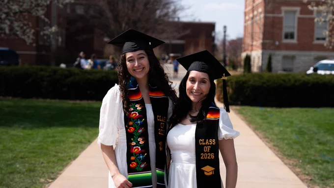 Karina and Ariana Hollis-Brau stand side by side on the Purdue University campus wearing black graduation caps and colorful embroidered stoles reading "Class of 2026."