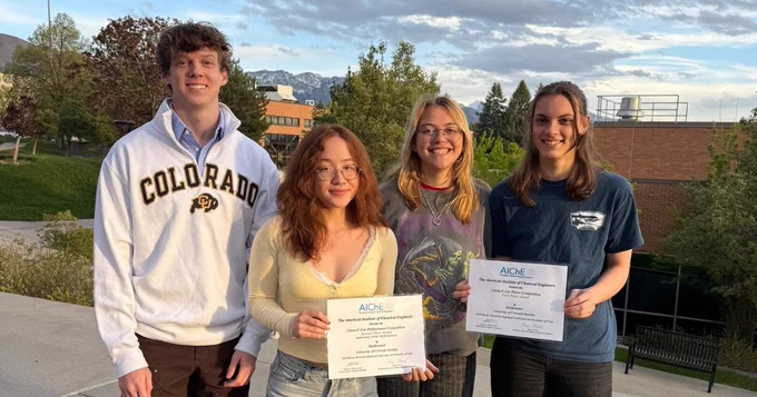 Four individuals are smiling and standing outdoors, with one holding Colorado State University certificates. They are positioned in front of a scenic background featuring trees, a building, and distant hills under a partly cloudy sky.