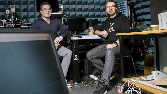 Two individuals are sitting in a tech laboratory, surrounded by electronic equipment. They are casually dressed, with one wearing a hoodie that has "KNL" printed on it. The room has a visible workstation with wires and a partially visible computer screen in the foreground.