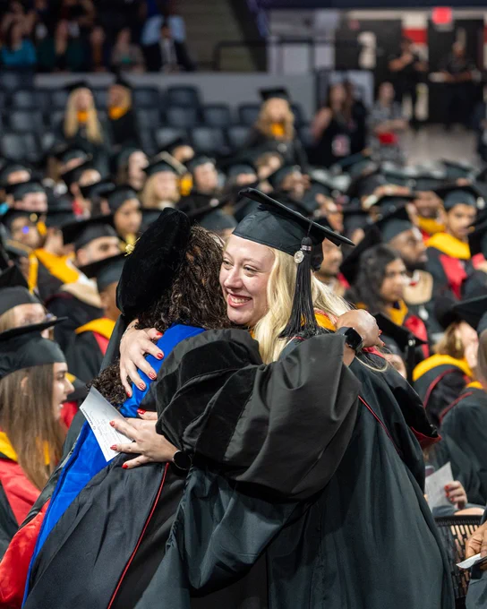 Two graduates in black caps and gowns embrace during a crowded commencement ceremony with seated attendees in the background.