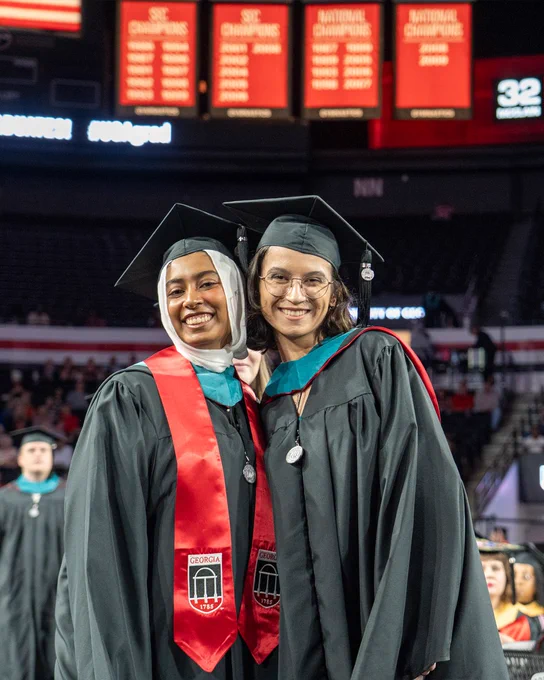 Two graduates in black gowns and red stoles with Georgia logos pose together at a commencement ceremony inside a large arena.