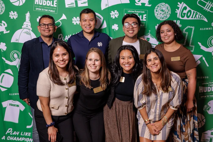 A diverse group of eight people poses together in front of a green backdrop decorated with sports-related graphics and phrases. They are smiling and appear to be at a social or professional event.