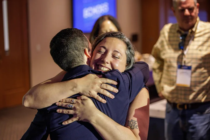 In a conference setting, two individuals share a heartfelt hug while another person stands close by, highlighting a moment of connection.