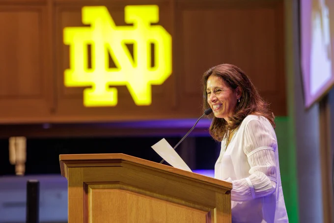 A woman stands at a podium, smiling as she speaks into a microphone. A large illuminated Notre Dame logo is visible in the background.