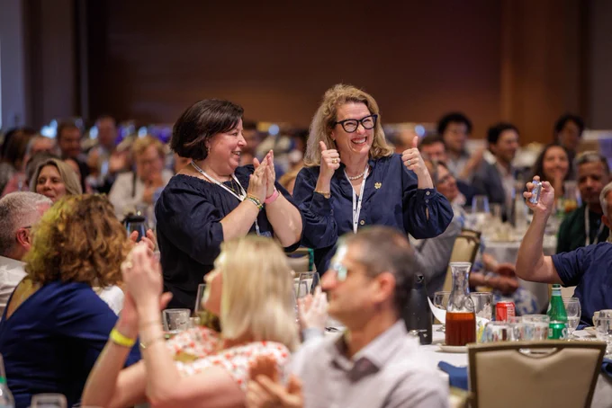 Two women stand in the center of a crowded room filled with seated attendees. One woman claps while the other gives a thumbs-up gesture, both smiling and engaging with the audience.