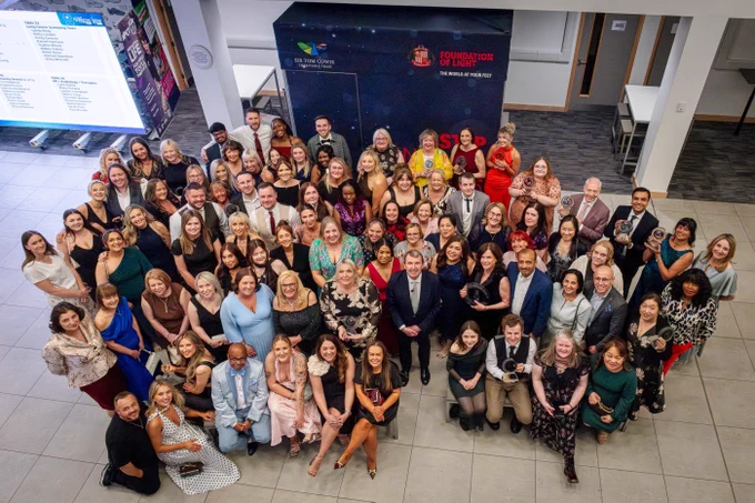 A large group of people are gathered together and smiling for a photo in a lobby area. Some wear formal attire. The backdrop features logos and text, including "The Prince's Trust" and "Foundation of Light.