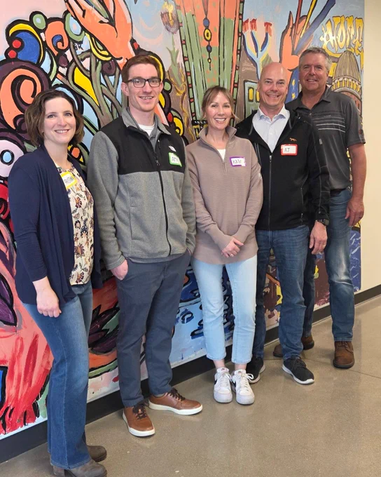 Five casually dressed adults standing in front of a colorful abstract mural inside a building hallway.