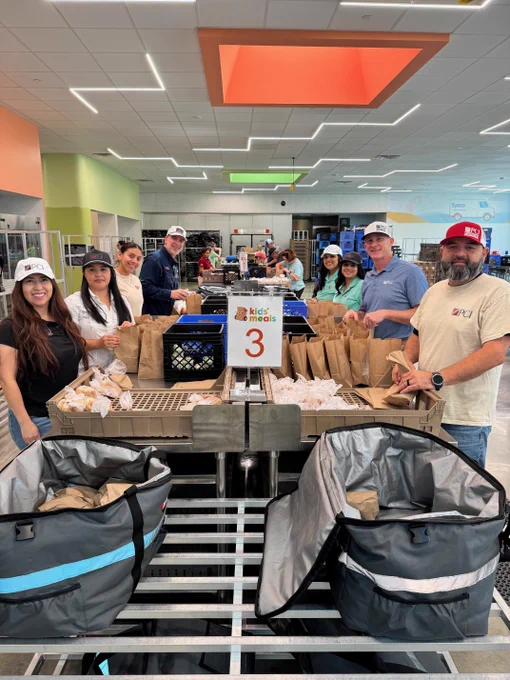Volunteers packing brown paper bags at a Kids Meals assembly station numbered 3 in a bright community center.
