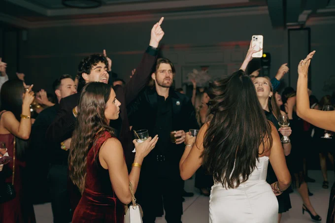 A lively gathering of elegantly dressed people in a dimly lit ballroom setting. The mood is festive and vibrant, with attendees holding drinks and capturing moments on their phones.
