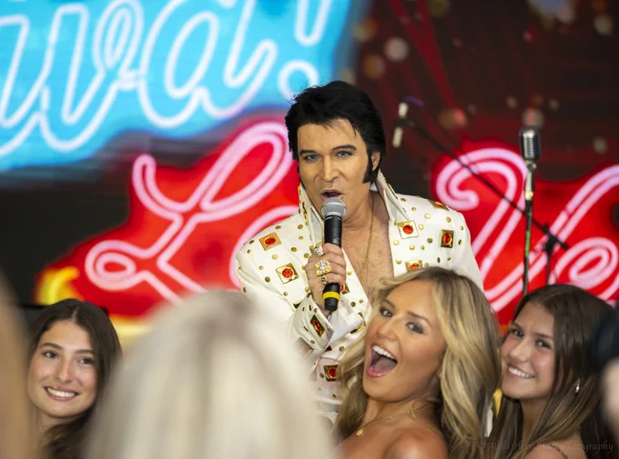 An Elvis impersonator performs enthusiastically in front of an audience with a vibrant neon backdrop. A group of smiling young women surround him.