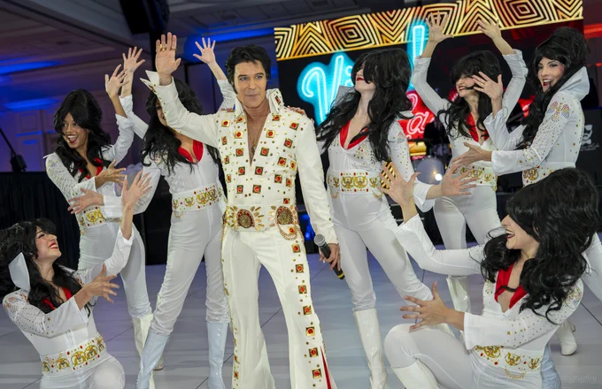 A lively performance featuring a person dressed as Elvis Presley, surrounded by dancers in similar costumes. The scene is vibrant and reminiscent of a retro Vegas show.