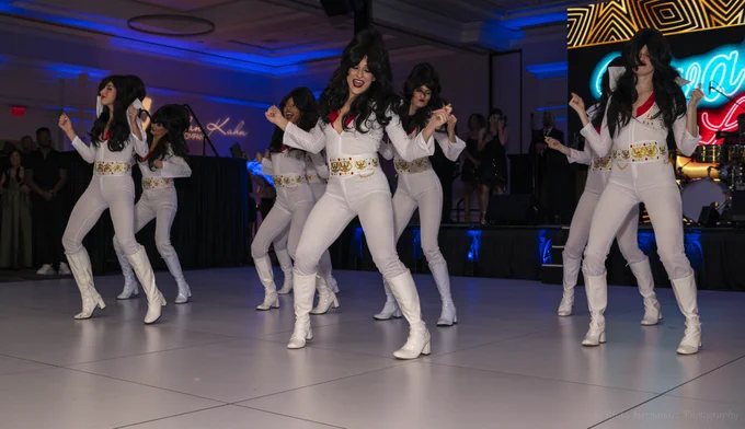 A group of performers dressed in Elvis Presley-inspired white jumpsuits and wigs dance energetically under colorful stage lights. The setting is lively and vibrant, indicative of a celebratory or themed event.