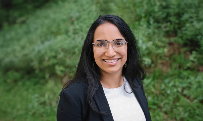 Headshot of Aparna Bhaduri smiling outdoors, wearing geometric glasses and a black blazer over a white top, with green foliage in the background.