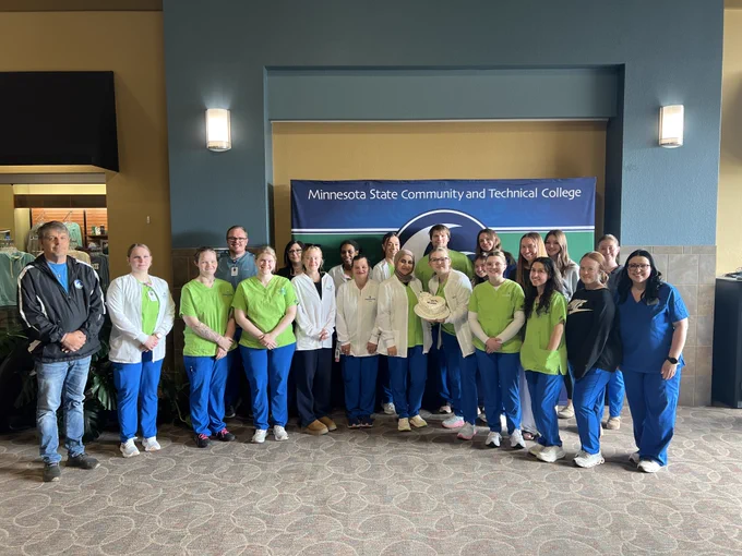 A group of people, some in medical or dental scrubs, stand together in front of a "Minnesota State Community and Technical College" banner. They are inside a building with patterned carpet and soft lighting.