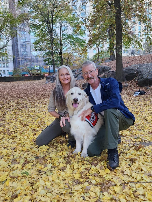 Joey, a woman with long gray hair, wearing a tan shirt and OD Green pants is smiling while kneeling next to Sampson, a fluffy Golden retriever wearing a red service dog vest, he is sitting in fall leaves in a park. On the other side is Jim, a smiling man who is kneeling with his arm around Sampson and his hand on Joey's knee. He is wearing OD green pants, a white t-shirt and Blue long sleeve over shirt. 