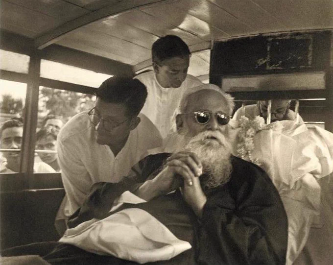 Rabindranath Tagore seated and being attended to by two people in a vehicle. There is a crowd visible through the window.