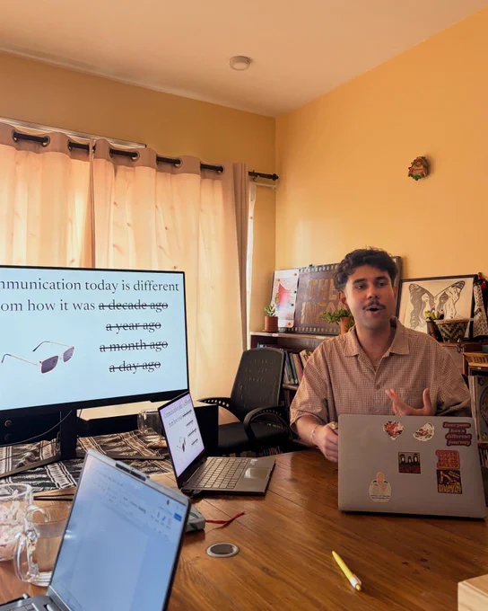 Image of a person who seems to be speaking as they type on their laptop. There is a monitor on the left side of the person. There are curtains and some paintings on the background.