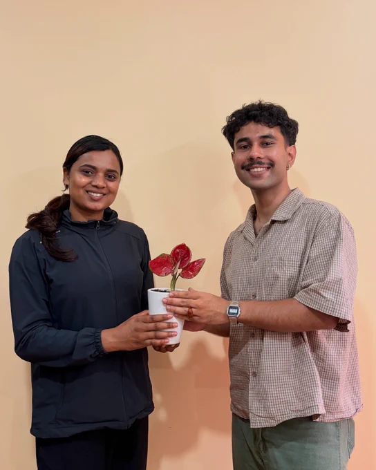 Two persons stand against a beige background. The person on the left is wearing a black jacket and handing over a white pot with plant with read leaves to the person on the right. The person on the right is wearing a shirt and a watch on their hands.