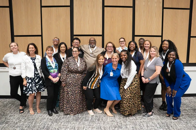 A group of teacher apprentices are standing together indoors, smiling at the camera. They are wearing a mix of professional clothing. The background features a wall with wood paneling and geometric design.