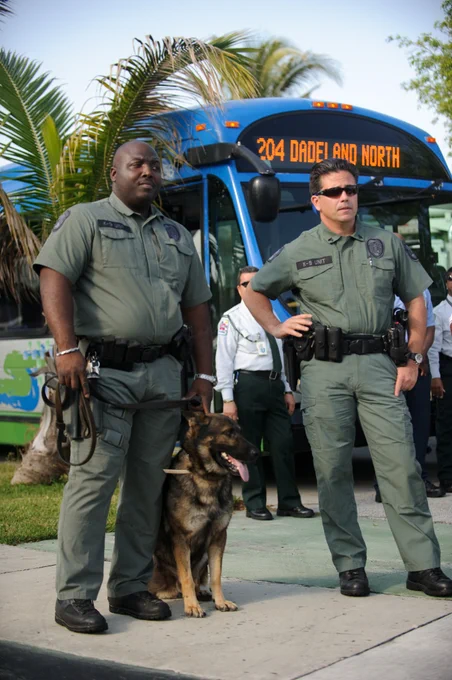 Two K-9 unit officers in khaki uniforms stand with a German Shepherd in front of a bus with \"204 DADELAND AND NORTH\" displayed. One officer holds the dog's leash. Others stand in the background, with palm trees visible.