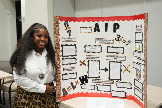 A person stands beside a presentation board titled "AIP." The board features various charts, text boxes, and graphics. The person is smiling and wearing a white shirt with a lanyard. The poster is decorated with a leopard print border.