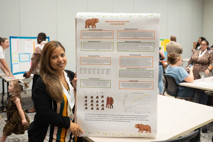 A person stands next to a presentation board featuring charts and images of brown bears. The background shows people gathering around tables with more posters and displays.