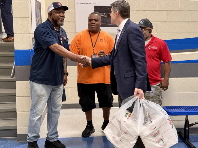 Treasurer Briner in a suit is shaking hands with one of three casually dressed teachers. Two of them are wearing badges and branded clothing. The person in the suit is holding several bags of Krispy Kreme donuts. They are standing in a hallway with a blue bench and staircase.