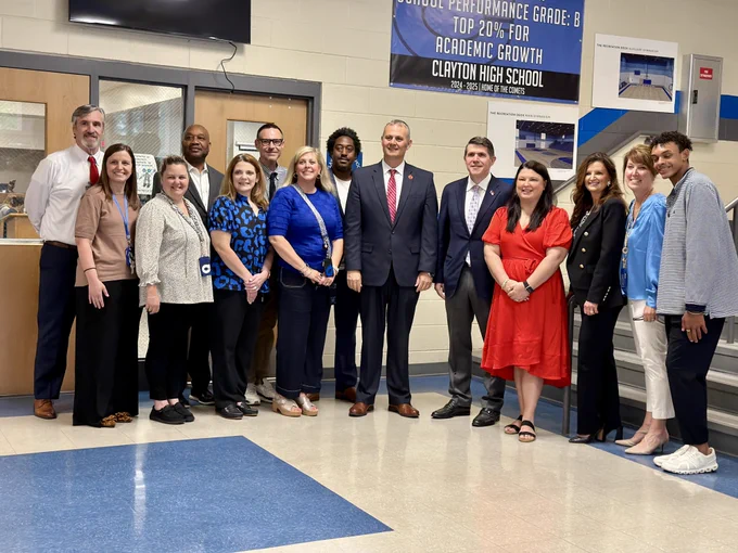 A group of individuals stand together, smiling, in a school hallway. They are dressed in business and casual attire. A sign in the background reads, "School Performance Grade: B Top 20% for Academic Growth Clayton High School.