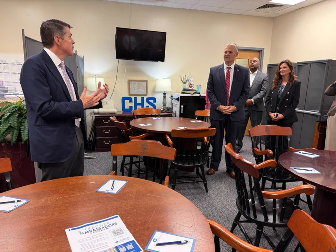 A group of school superintendents and Treasurer Briner standing in a room with a "CHS" sign on a shelf. A person speaks while others listen, with tables and chairs arranged in the foreground. Pens and papers are placed on the tables. A television is mounted on the wall.