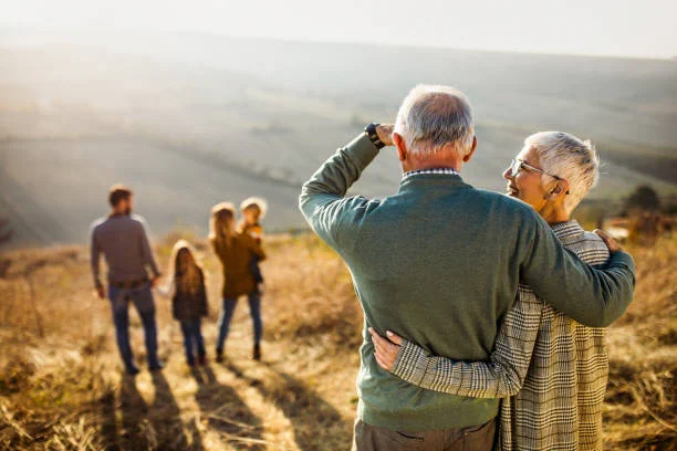 Elderly couple embracing and looking out over a sunlit countryside with a younger family walking ahead on a dirt path.