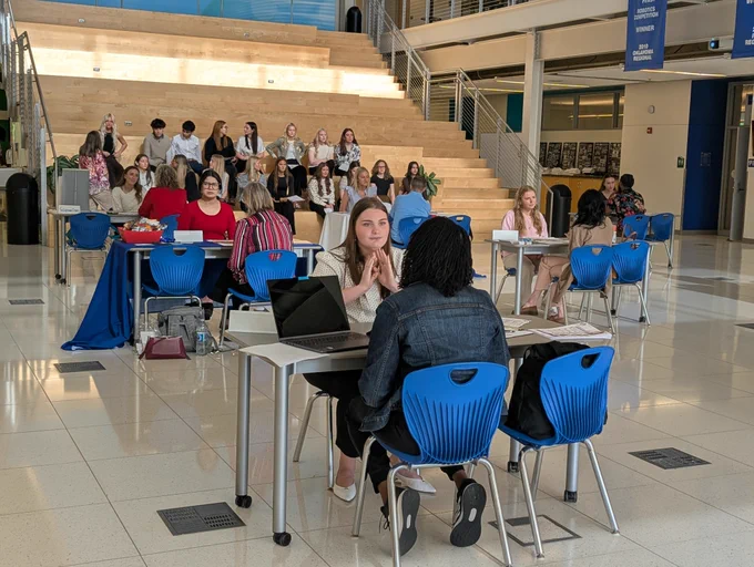 A group of people, mostly students, seated at tables in a school-like setting, engaged in various activities like using laptops, discussing, and interviewing.