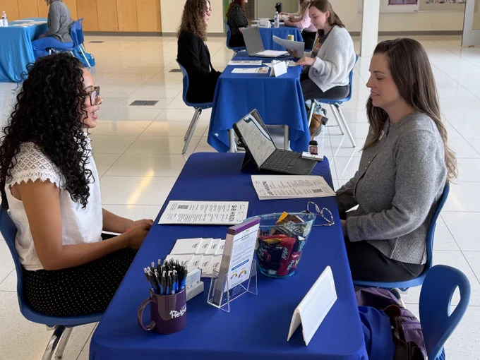 Several students working at desks in a classroom setting. Blue tables, various laptops and supplies visible. A young woman interviewing with a healthcare professional at a table in an atrium of CAPS.