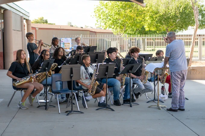 A group of young musicians playing saxophones and clarinets outdoors while a conductor leads them during a performance