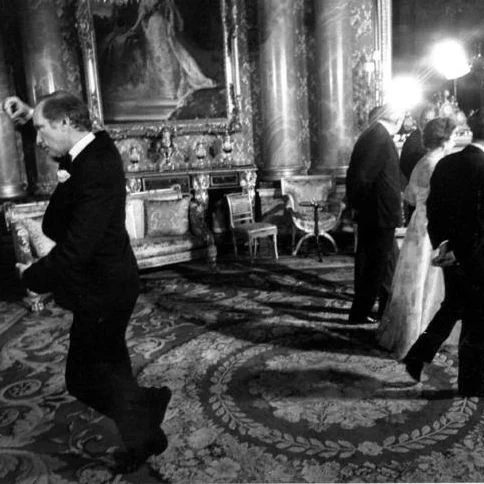 Pierre Trudeau in a formal interior setting, shown walking across an ornate room with several other people in formal attire. He wears a dark suit and appears mid-step, with one arm slightly raised. The room features patterned carpets, tall decorative columns, and framed artwork on the walls. Other individuals stand and converse in the background under bright interior lighting.
