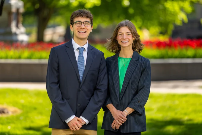 Two people are standing outdoors in a park with vibrant green grass and red flowers in the background. Both are smiling and dressed in business attire.