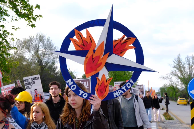 NATO amblemi üzerine alev çizimleri eklenmiş “No to NATO” yazılı büyük bir döviz taşınıyor. Arkada yürüyüş korteji ve çeşitli pankartlar görülüyor.