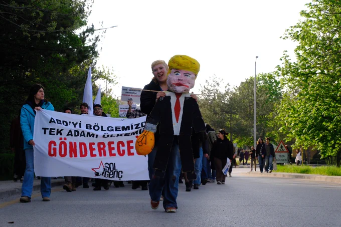 bir öğrenci Donald Trump maketiyle yürüyüş sırasında poz veriyor. Kalabalık arasında gülümseyen ve slogan atan öğrenciler dikkat çekiyor.