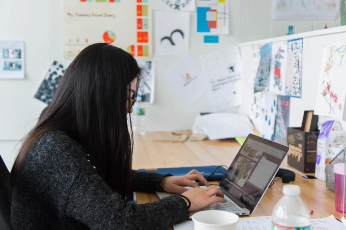 Person with long dark hair types on a laptop at a cluttered desk with printed designs pinned to the wall and a coffee mug nearby.\n