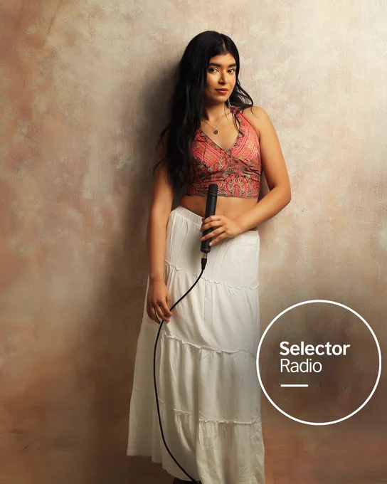 Shivani Sen, a woman in a patterned halterneck crop top and long white skirt holds a microphone against a textured warm studio backdrop. The Selector Radio logo appears in a white circle in the bottom right corner.