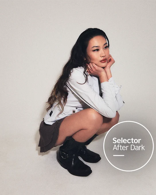 Producer Baobei, a woman with long dark hair crouches thoughtfully in black boots, black mini skirt and a white jacket against a plain white studio background. The Selector After Dark logo sits in a white circle at the bottom right.