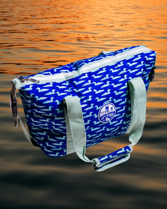 A blue tote bag with a white bird pattern and a logo, set against a sunset water background. The image has a relaxed and adventurous mood.