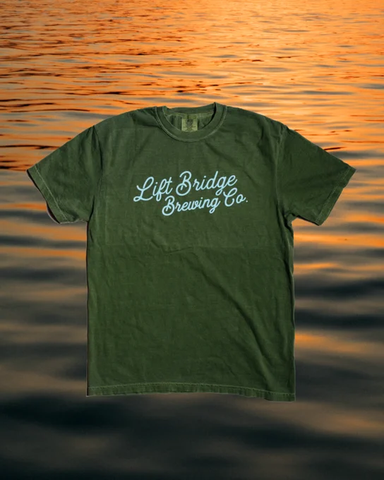 A green t-shirt featuring the Lift Bridge Brewing Co. logo placed against a serene water background at sunset, evoking a calm and relaxed mood.