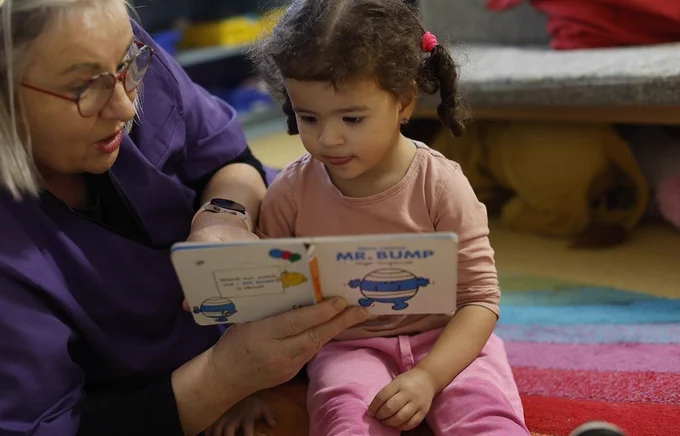 Photograph shows an early years educator lying on the ground, reading a book to a young child, who is sitting on the floor beside her.