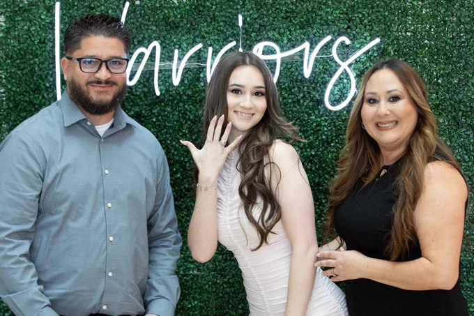 Three people are posing in front of a green, leafy backdrop with the word "warriors" in bright script. The person in the center is displaying a ring on one hand, smiling. The person on the left has a beard and glasses. The person on the right has long, styled hair and is smiling.