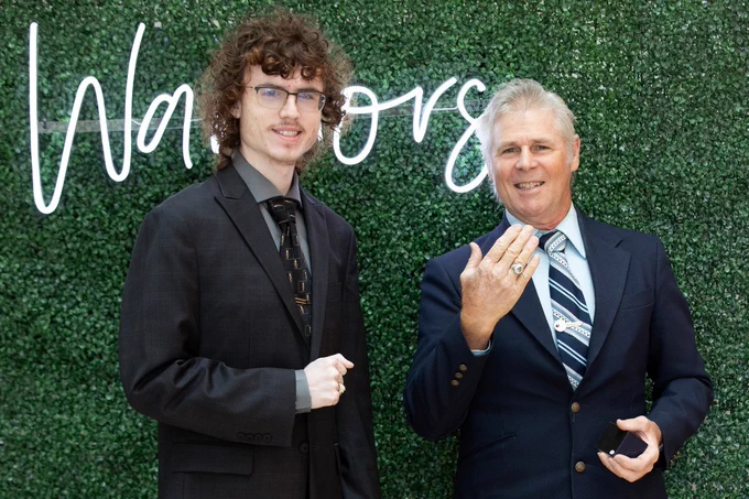 Two individuals stand in front of a hedge wall with "Warriors" written in script. Both are wearing suits, and one is displaying rings on their fingers.