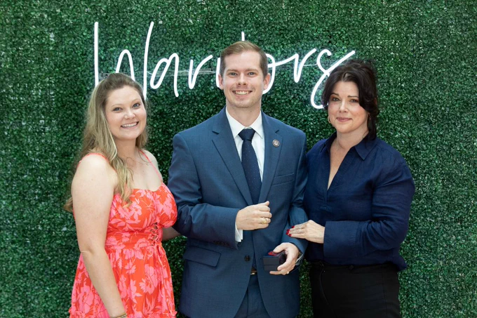 Three people are smiling in front of a green leafy backdrop with the word "Warriors" written in cursive. The individual in the middle is wearing a suit, while the others are dressed in vibrant clothing.