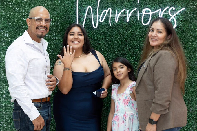 A group of four people smiling in front of a green foliage wall with the word "Warriors" in white script. One person is holding up a hand to show a piece of jewelry. A child is among the group, wearing a floral dress.