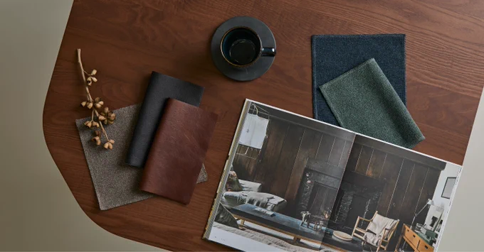 Wooden table with fabric swatches, a cup of coffee, dried flowers, and an open magazine showing a cozy living room interior.