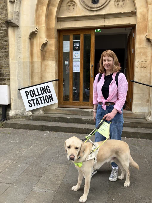 Otto, a blond Labrador golden retriever cross  guide dog standing on a stone pavement outside a building which is being used as a polling station.  I am standing next to him. I am  smiling, facing the camera, and wearing a black T‑shirt, a light pink long-sleeve shirt tied at the waist, loose blue jeans, silver trainers, and a backpack with dark straps.  
Behind us is a doorway with wooden-framed glass doors and stonework around the entrance.  
To the left of the doorway, a large white rectangular sign hangs at an angle, reading “POLLING STATION” in bold black capital letters.
